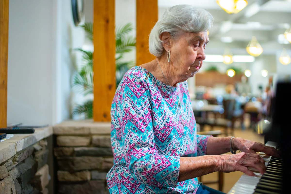 Elderly woman playing piano in a communal setting.
