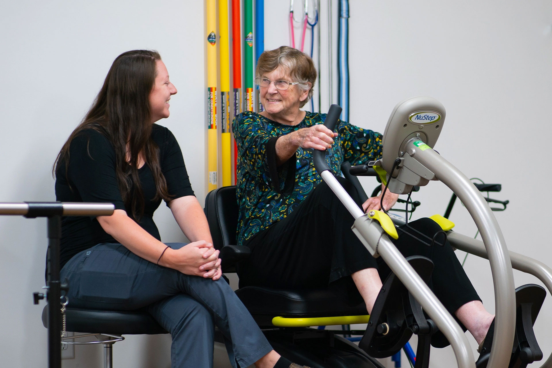 Two women conversing in a rehabilitation setting, one exercising.